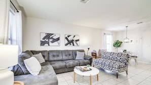 A modern living room with a grey sectional sofa, a patterned armchair, and a white coffee table. There are abstract paintings on the wall and a dining area in the background. - Visit Route 66! Pet-friendly Joplin Gem w/ Yard (Joplin)