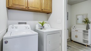 A laundry area with two white front-loading washers and dryers, wooden cabinets above, and a white vanity with a mirror and shelves. - Visit Route 66! Pet-friendly Joplin Gem w/ Yard (Joplin)