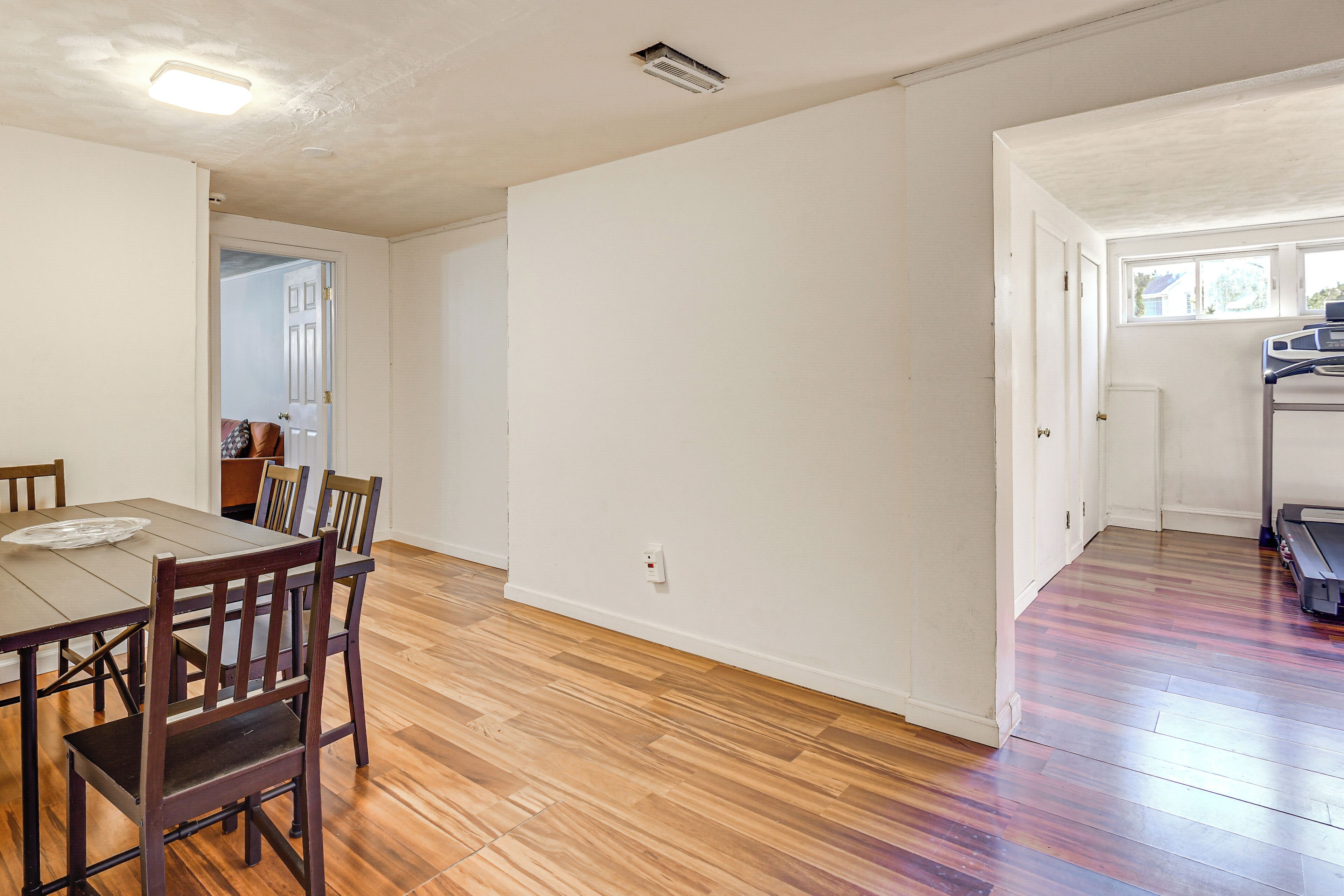 A dining area with a wooden table and chairs, a kitchen area with a refrigerator and a window, and a hallway leading to another room.