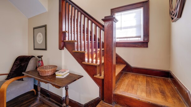 A wooden staircase with a handrail, a small table with a basket and books, a chair with a dark throw, and a framed picture on the wall. - Walk to Main St & Campus! Dtwn Radford Home (Radford)