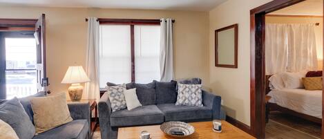 A living room with a grey sofa, a wooden coffee table, and a window with white curtains.