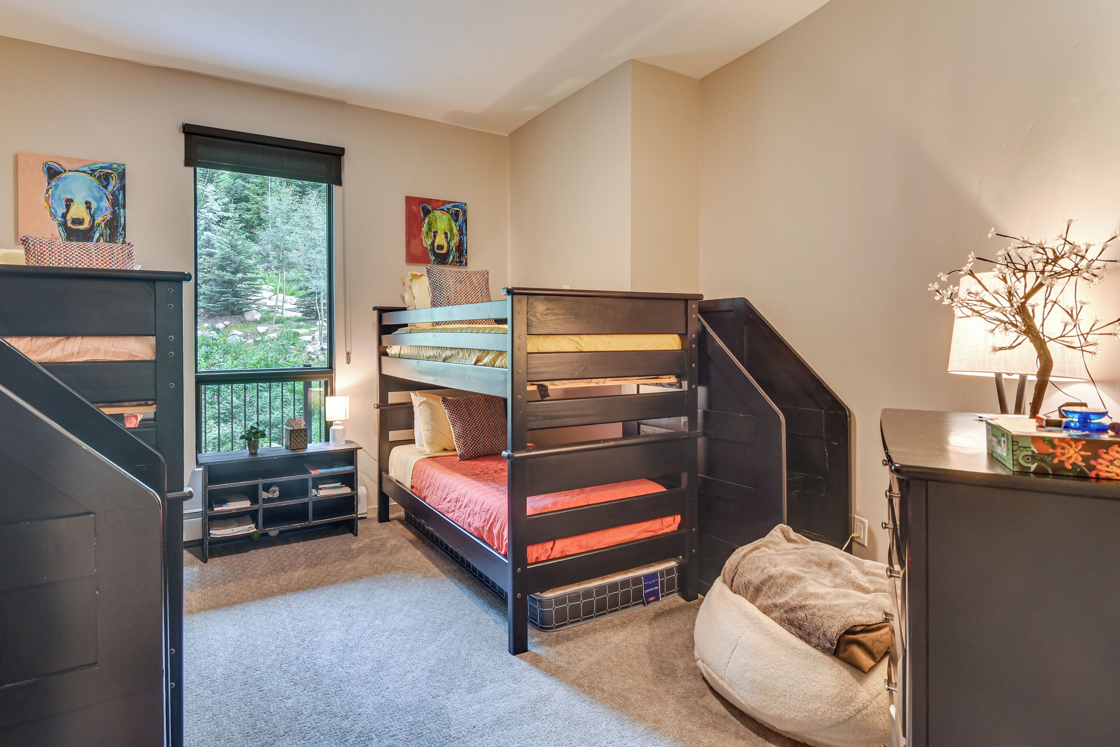 A bedroom with bunk beds, a window with a view of trees, and a dresser with a lamp.