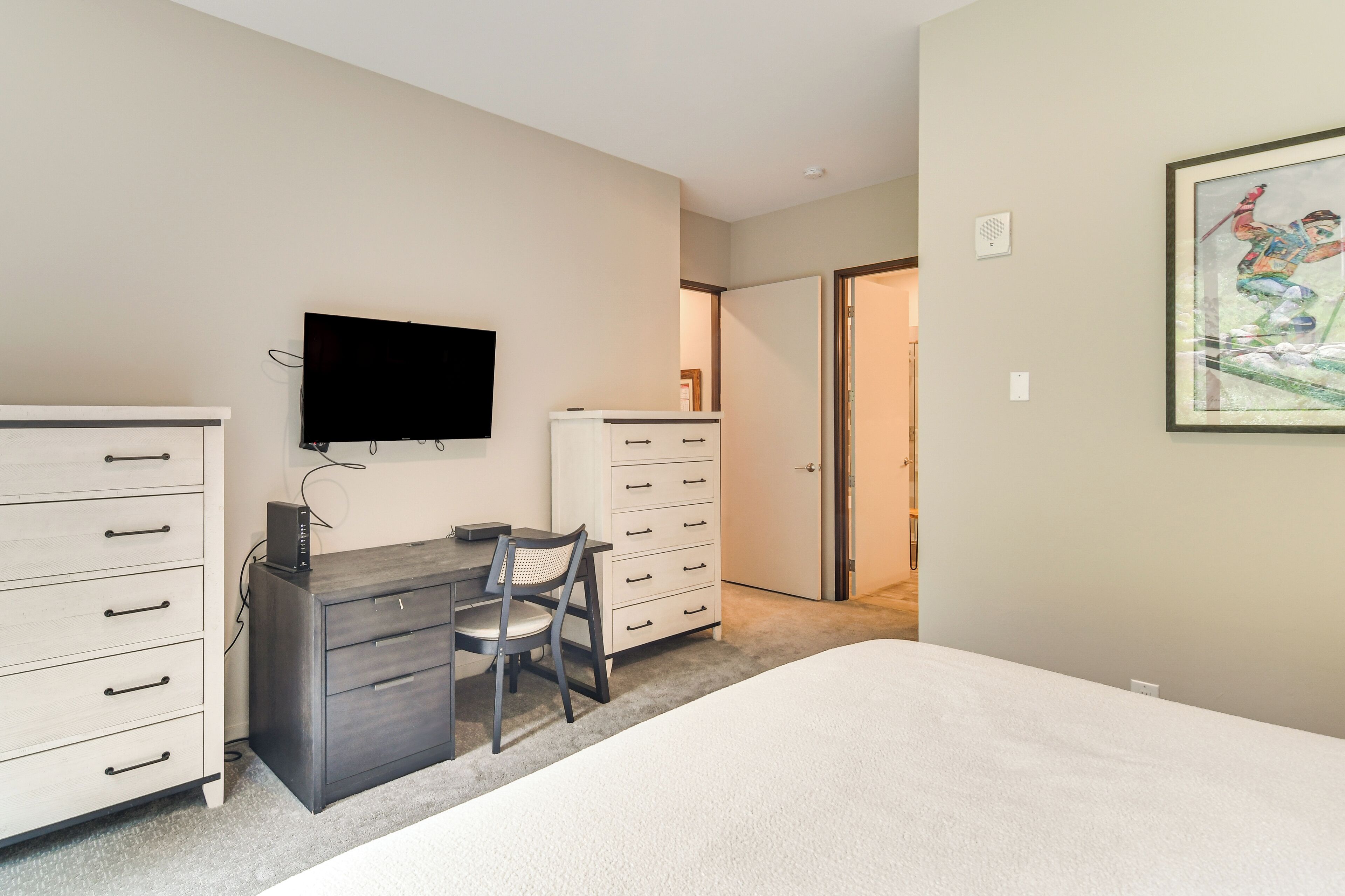 Un dormitorio con cama, escritorio y silla, cómoda y televisor en la pared.