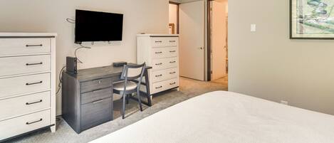 Un dormitorio con cama, escritorio y silla, cómoda y televisor en la pared.