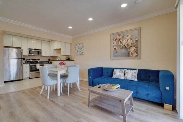 Una cocina moderna con armarios blancos, electrodomésticos de acero inoxidable y un área de comedor con un sofá azul y una mesa de centro.