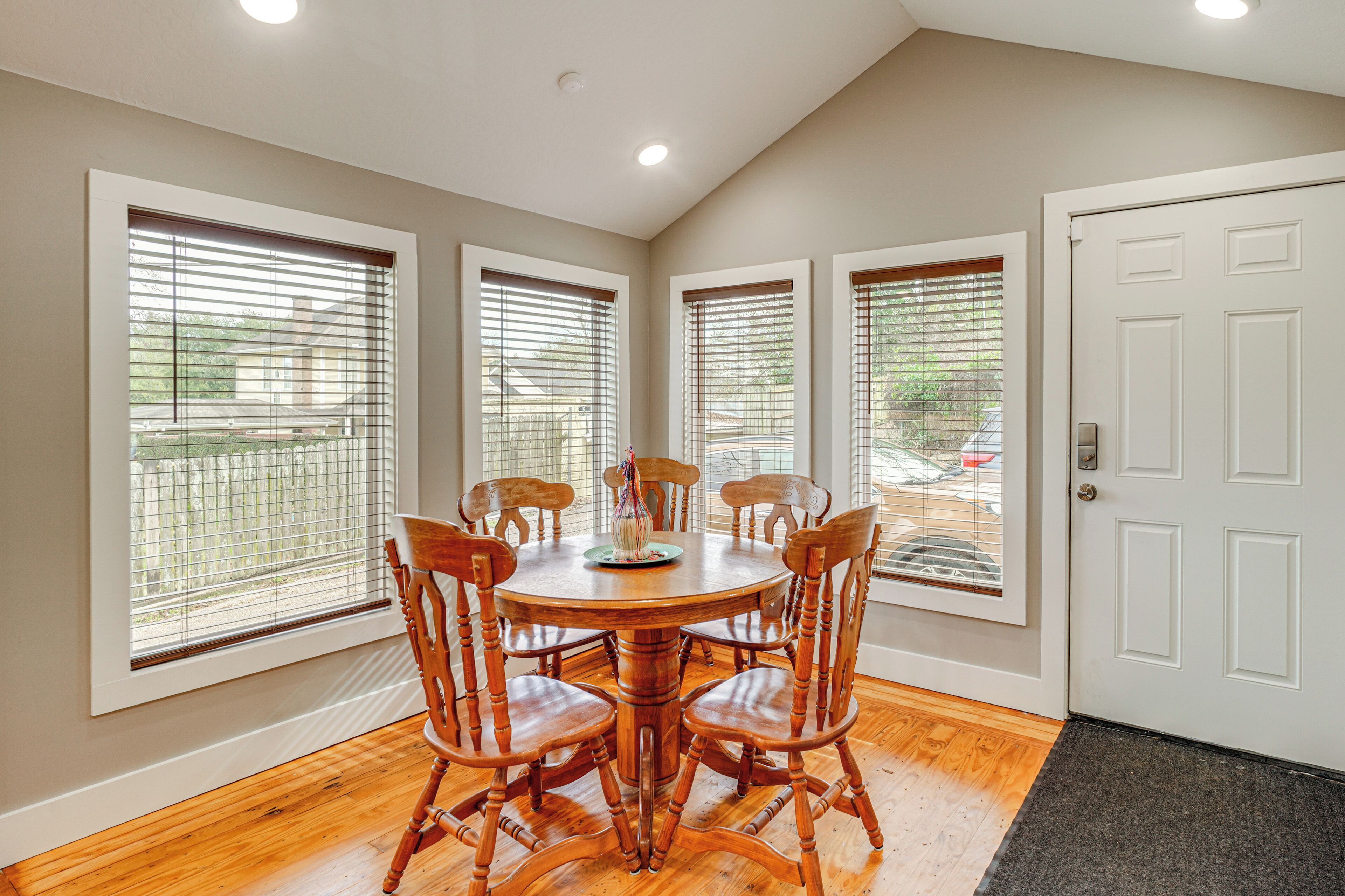 A dining area with a round wooden table surrounded by six wooden chairs, large windows with blinds, and a white door.