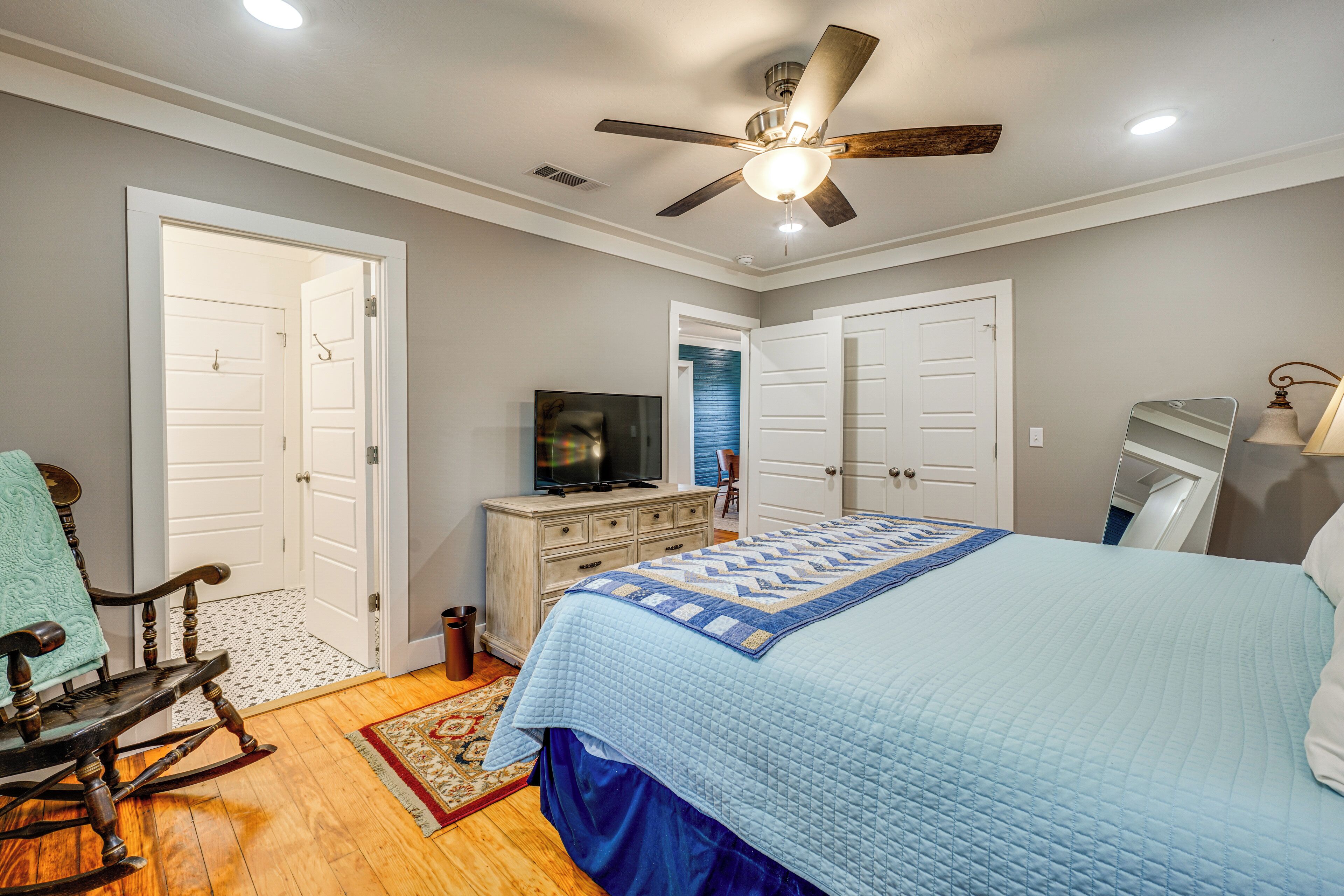 A bedroom with a bed, a rocking chair, a dresser, a television, and a ceiling fan.