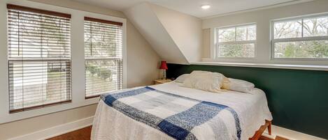 A bedroom with a large bed, wooden flooring, and multiple windows with blinds.
