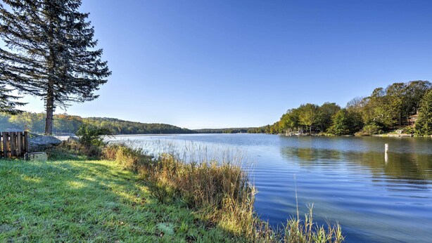 A serene lakeside view with a wooden dock, grassy banks, and a dense forest in the background. - Poconos Retreat in The Hideout Lake Community! (Lake Ariel)