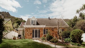 Uma casa de pedra com telhado de cerâmica, entrada em pedra e um jardim com diversas plantas e um caminho que leva à porta.