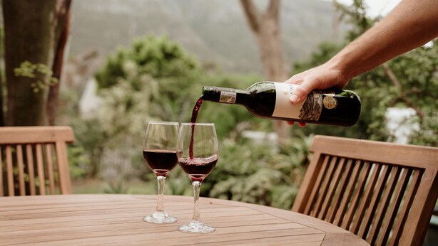 Uma mão servindo vinho em duas taças sobre uma mesa de madeira ao ar livre.