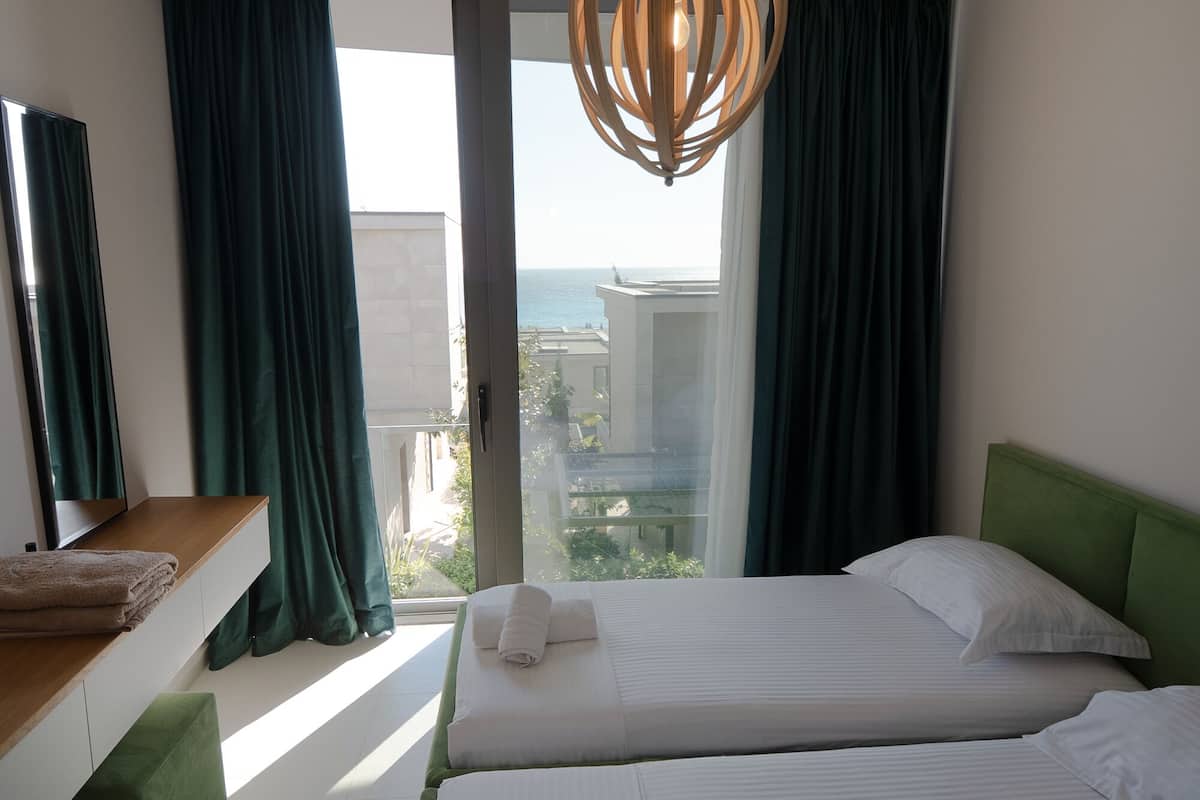 Habitación de hotel con cama, banqueta, otomana verde, lámpara colgante y vista al mar.