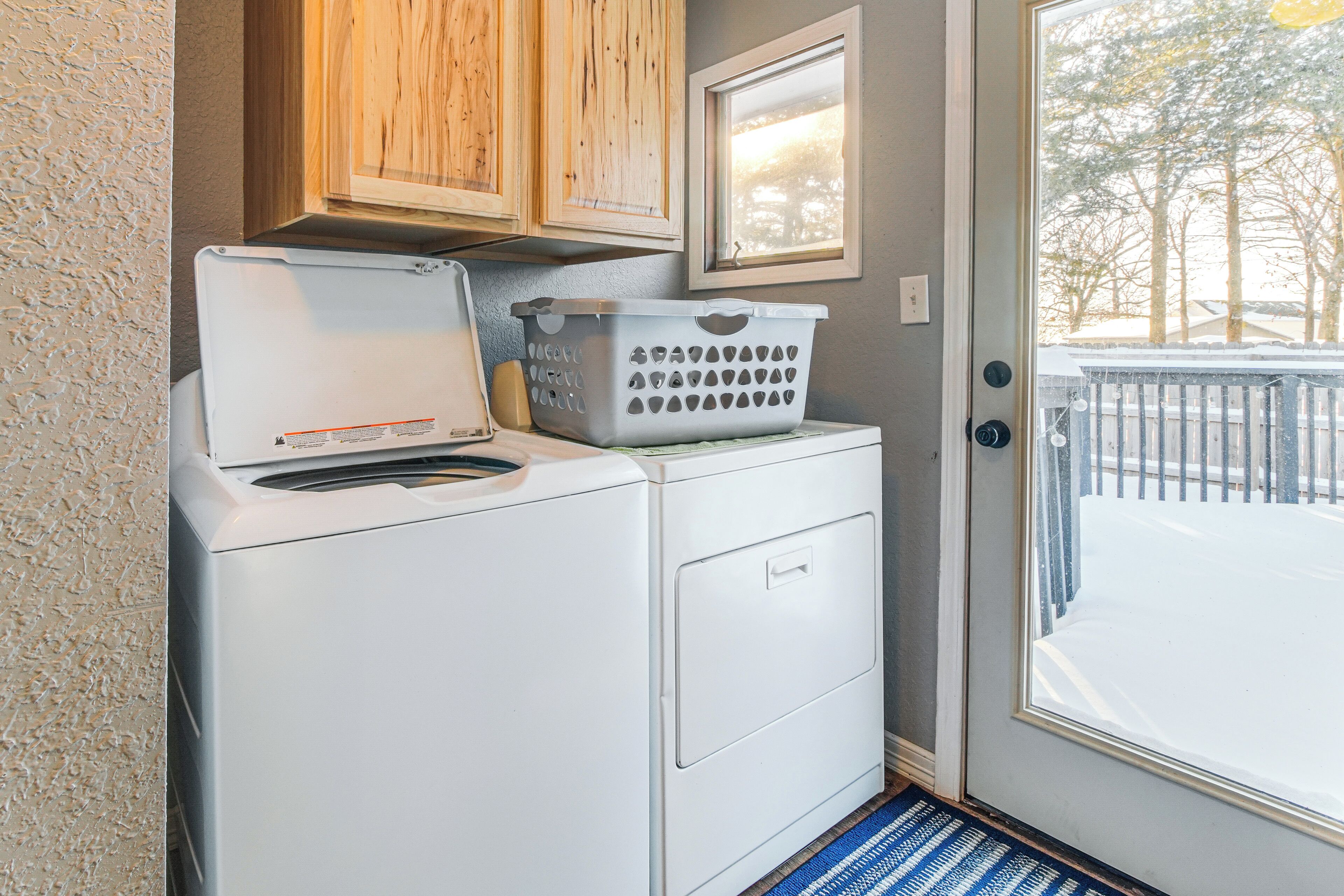A compact laundry area with a front-loading washer and dryer, a wooden cabinet, a framed picture, and a view of a snowy outdoor deck.