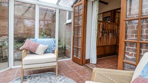 Une véranda avec une chaise en bois et un petit tapis, un mur en briques et une porte en bois donnant sur l’intérieur.
