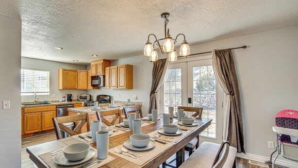 A dining area with a wooden table set for four, surrounded by wooden chairs. A chandelier hangs above, and a window with curtains is visible. - Family & Group Friendly! Hilltop Cedar City Home (Cedar City)