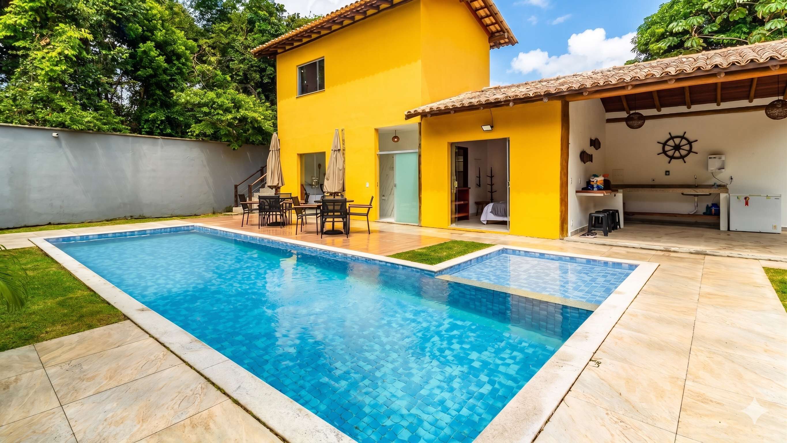 Una piscina con un borde de baldosas, una casa amarilla con techo de tejas y una pequeña zona de patio amueblada al aire libre.