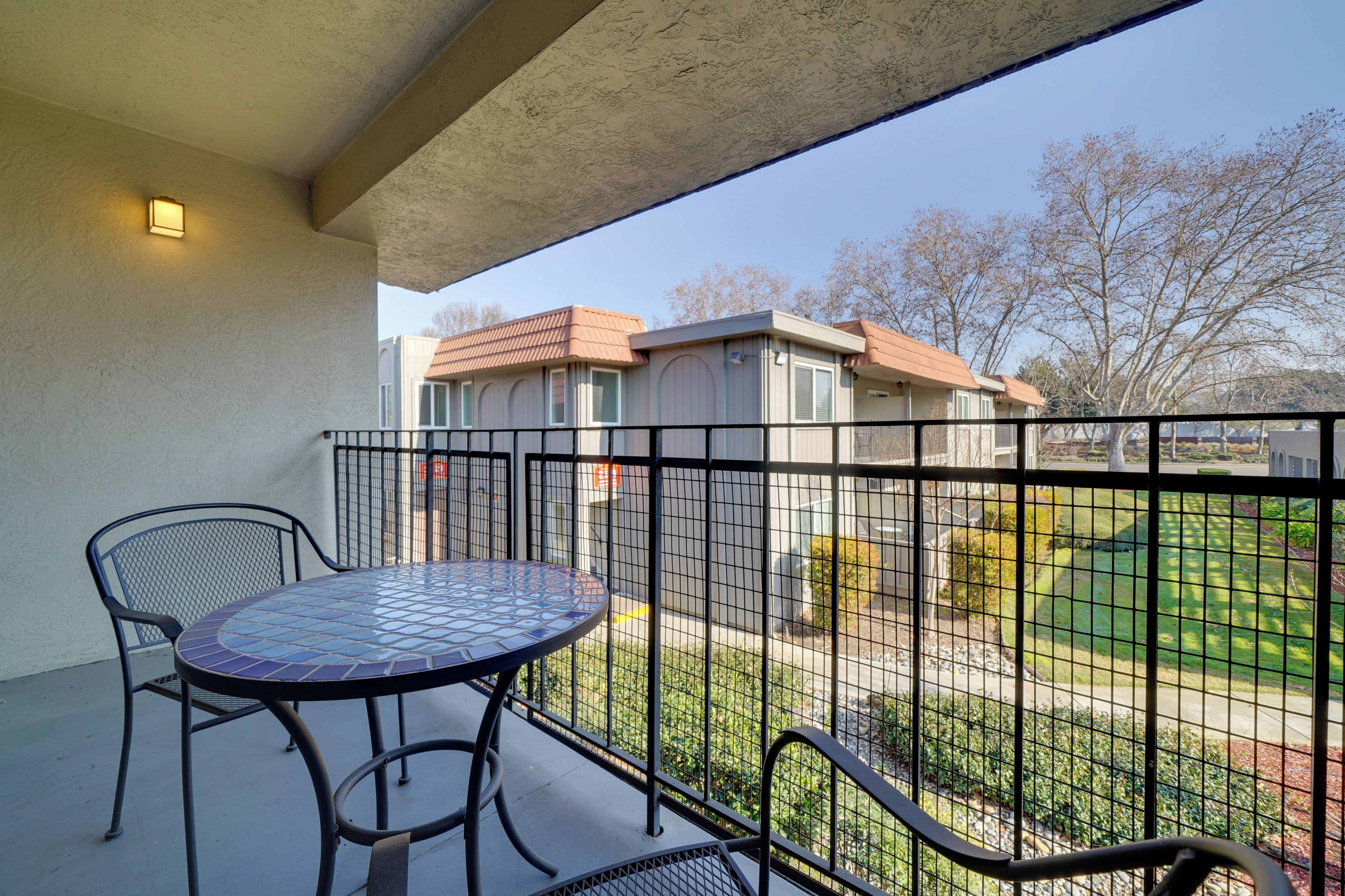 A balcony with a round table and two chairs, overlooking a residential area with houses and a lawn.