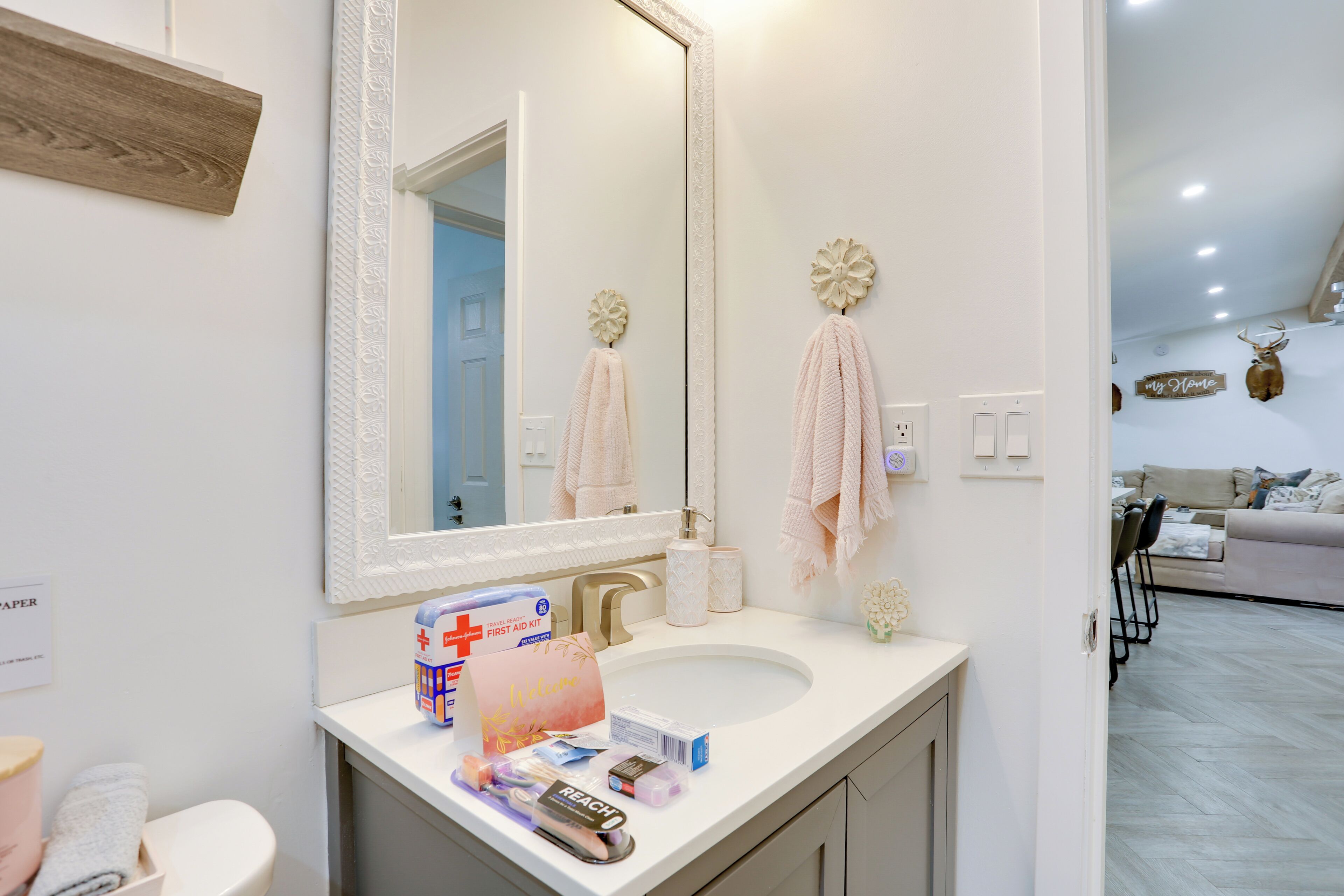 A bathroom with a large mirror, a sink, and a toilet. There are towels hanging on the wall, a first aid kit, and a welcome bag on the counter.