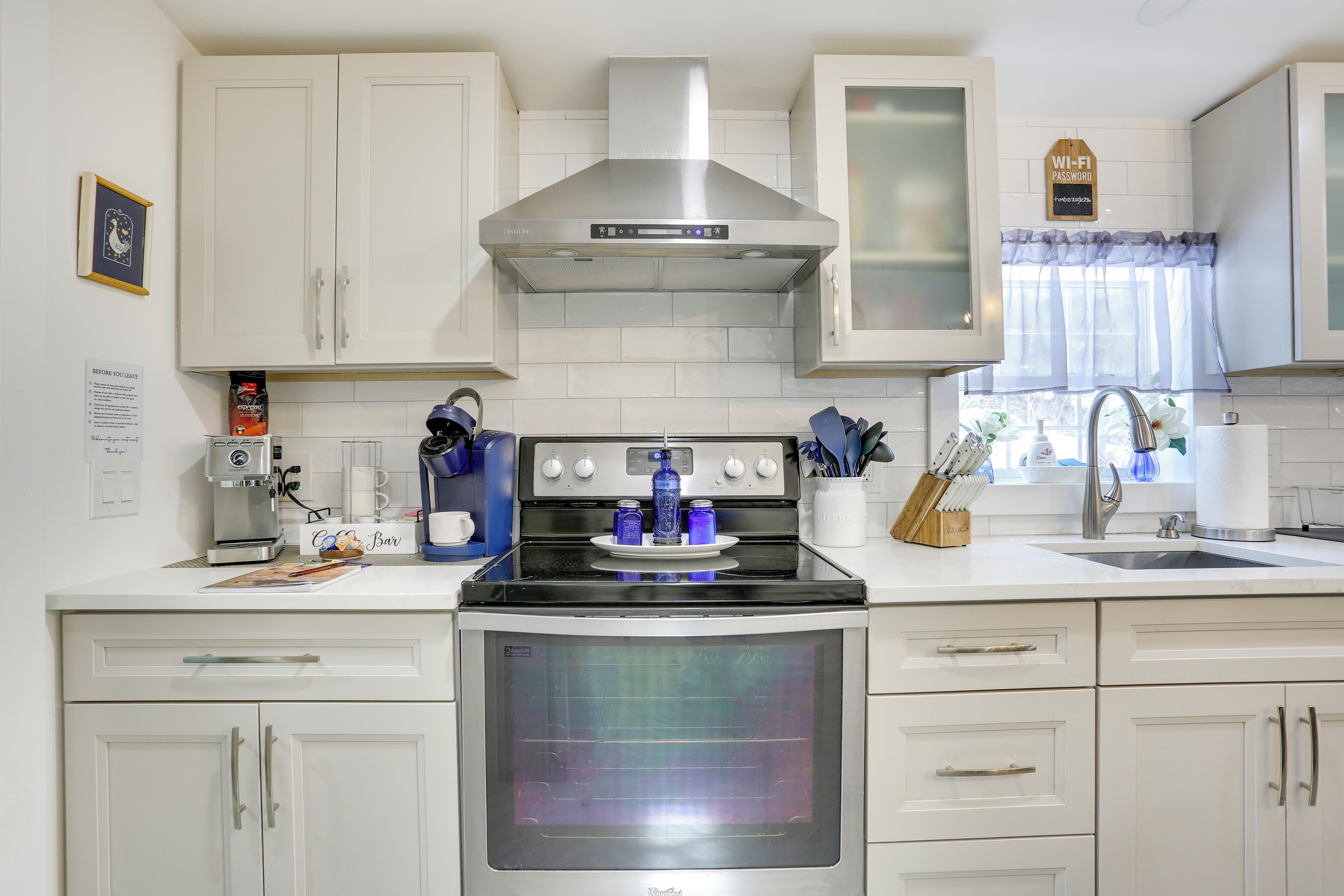 A kitchen with white cabinets, a stainless steel range hood, a built-in oven, and a coffee station.