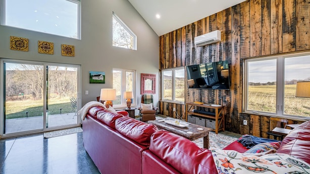 A spacious living room with a red leather sofa, a television mounted on a wooden wall, and large windows offering a view of the outdoors. - Peaceful Farmhouse Retreat 6 Mi to Virginia Tech! (Blacksburg)