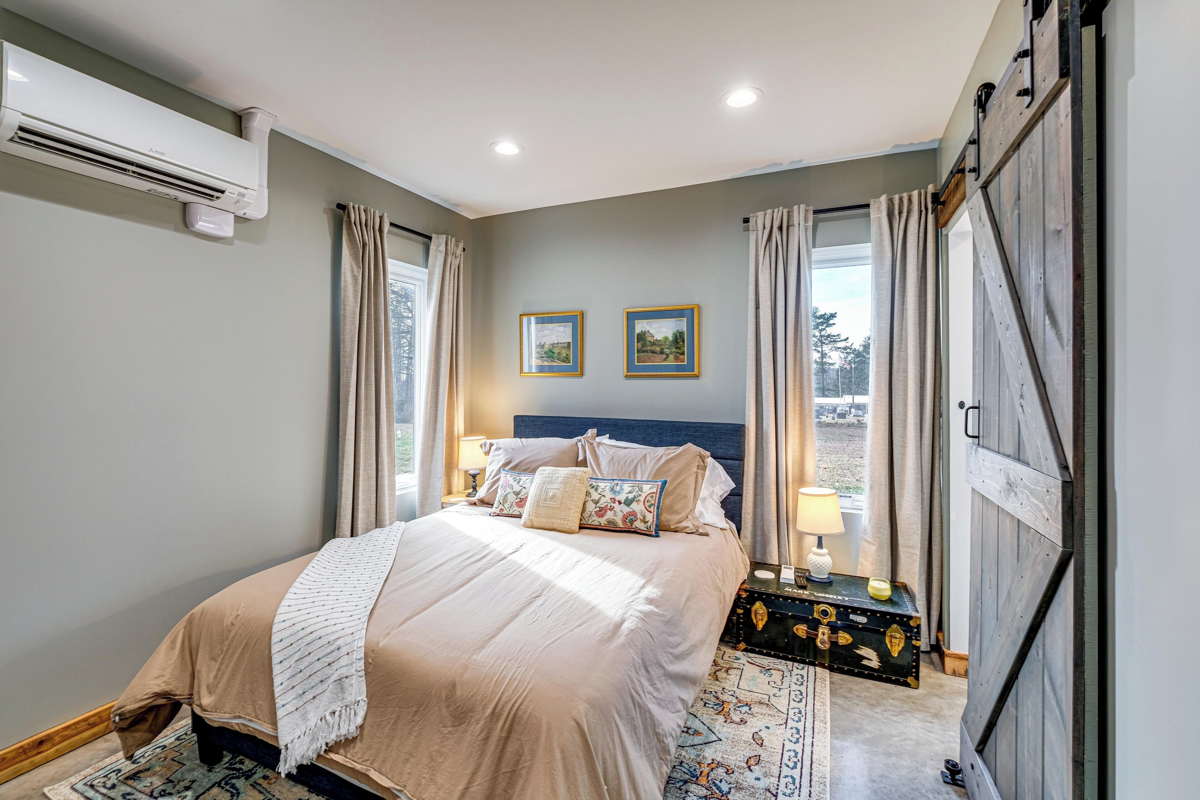 A bedroom with a bed, two framed pictures on the wall, a bedside table with a lamp, and a large window with curtains.