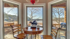 A dining area with a round table and four wooden chairs, large windows with a view of a deck and trees, and a red chandelier. - 2-tier Dock! Jordan Lake Family Retreat (Titus)