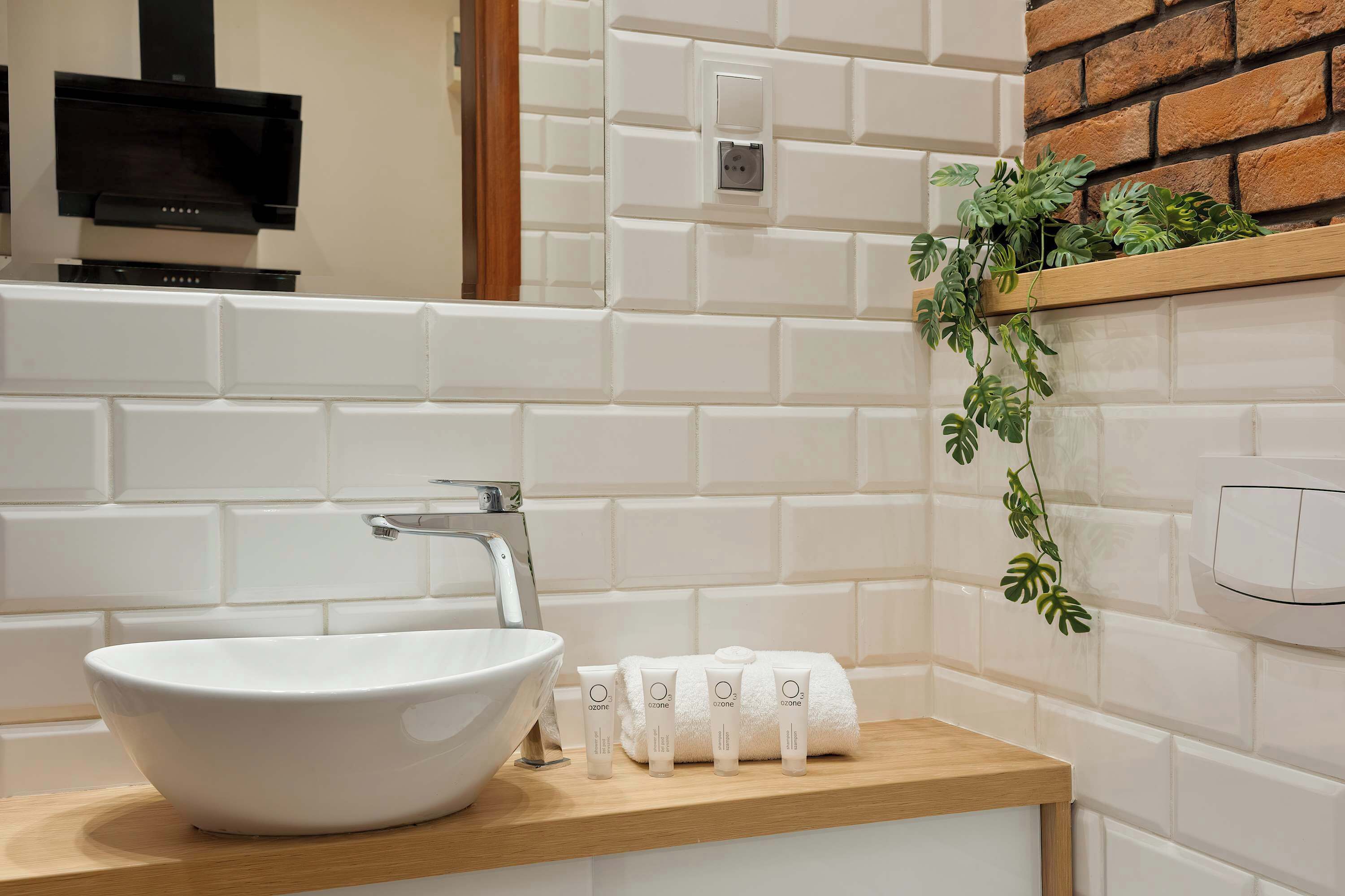 Ein Badezimmer mit einem weißen Waschbecken, einer Holzauslage, einem Spiegel, einer schwarzen Dunstabzugshaube, einer weißen Fliesenwand, einem Holzregal mit Pflanzen und einem weißen Handtuchspender.
