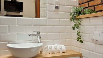 Ein Badezimmer mit einem weißen Waschbecken, einer Holzauslage, einem Spiegel, einer schwarzen Dunstabzugshaube, einer weißen Fliesenwand, einem Holzregal mit Pflanzen und einem weißen Handtuchspender.
