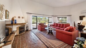 A living room with a fireplace, red sofa, and a large window with a view of greenery. - Casa Rouxinol in Carvoeiro (Lagoa)