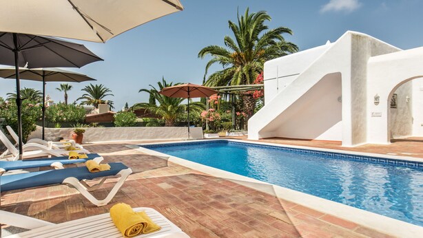 A swimming pool with lounge chairs and umbrellas, surrounded by palm trees and a clear sky. - Casa Rouxinol in Carvoeiro (Lagoa)