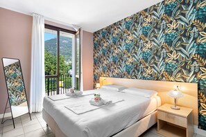 A hotel room with a bed, a bedside table with a lamp, a mirror, and a view of greenery outside. - Mobilehome in Porlezza am Luganersee (Porlezza)