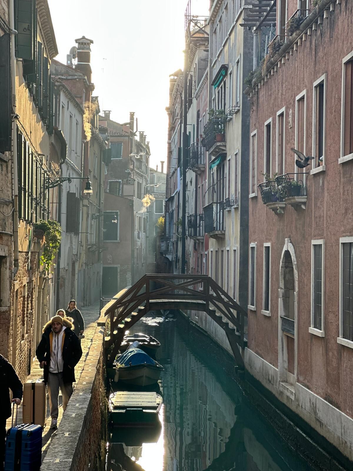 Een smalle gracht in een historische Europese stad, met gebouwen aan weerszijden, een houten brug en boten afgemeerd langs het water.
