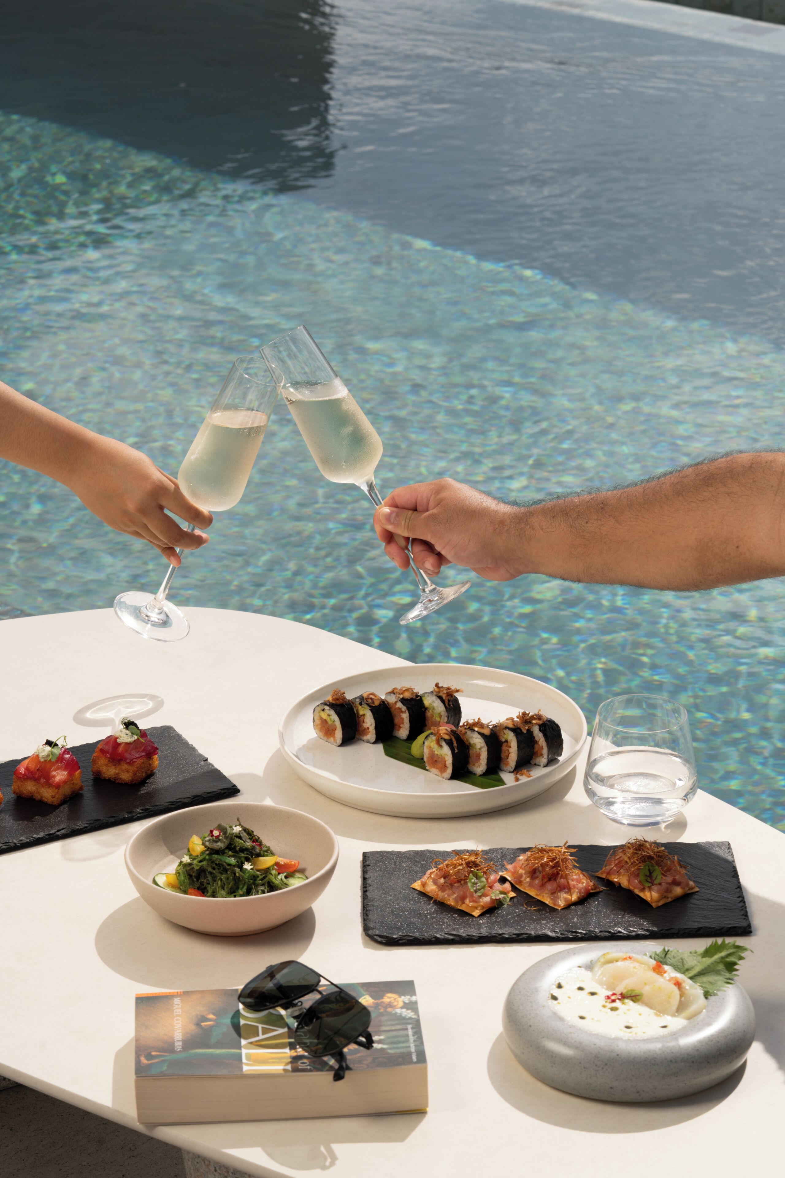 Una mesa con sushi, un tazón de ensalada, un libro y un vaso de agua.