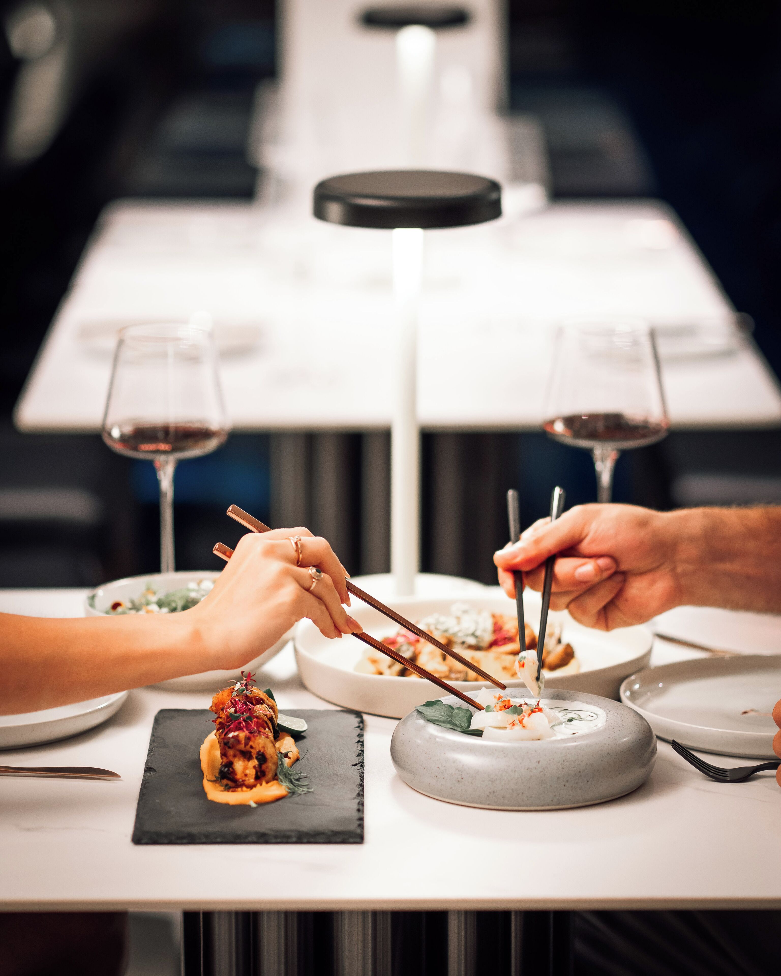 A dining table with plates of food, chopsticks, and wine glasses.