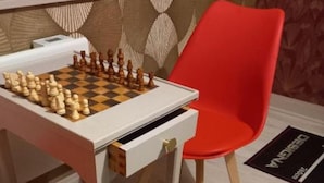 Un tablero de ajedrez sobre una mesa blanca con un cajón, una silla roja y un papel tapiz estampado al fondo.