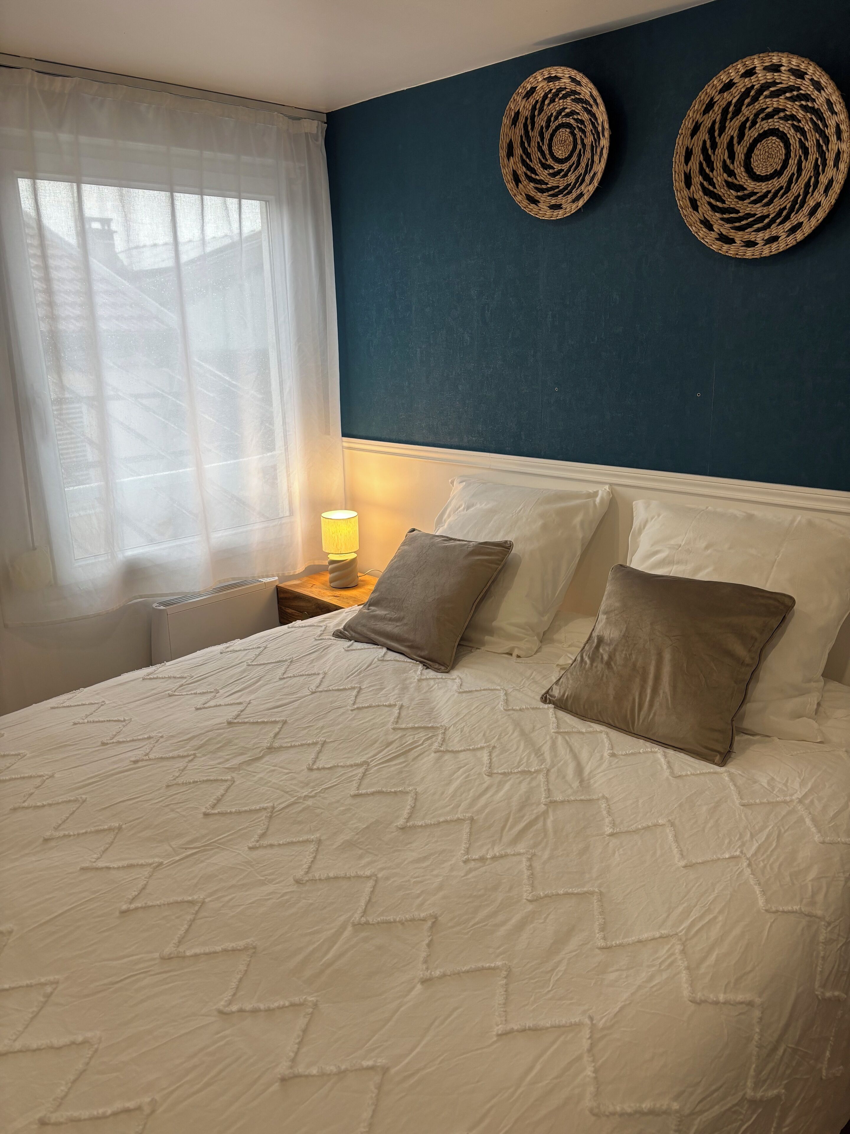 Un letto con una coperta a motivi, due decorazioni tessute appese al muro, una lampada da comodino e una finestra con tende leggere.