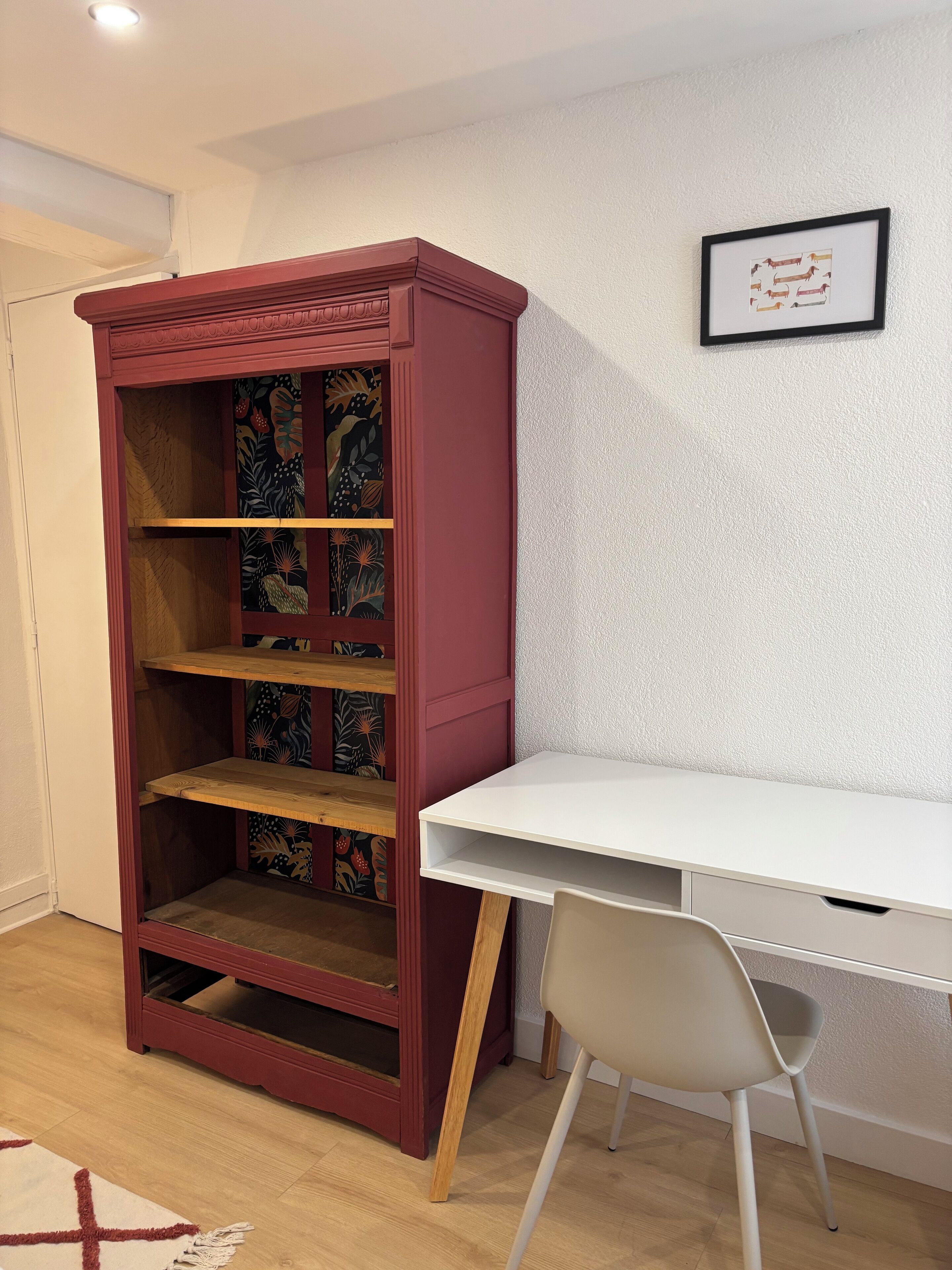 Una habitación con una estantería roja, un escritorio blanco y un cuadro en la pared.