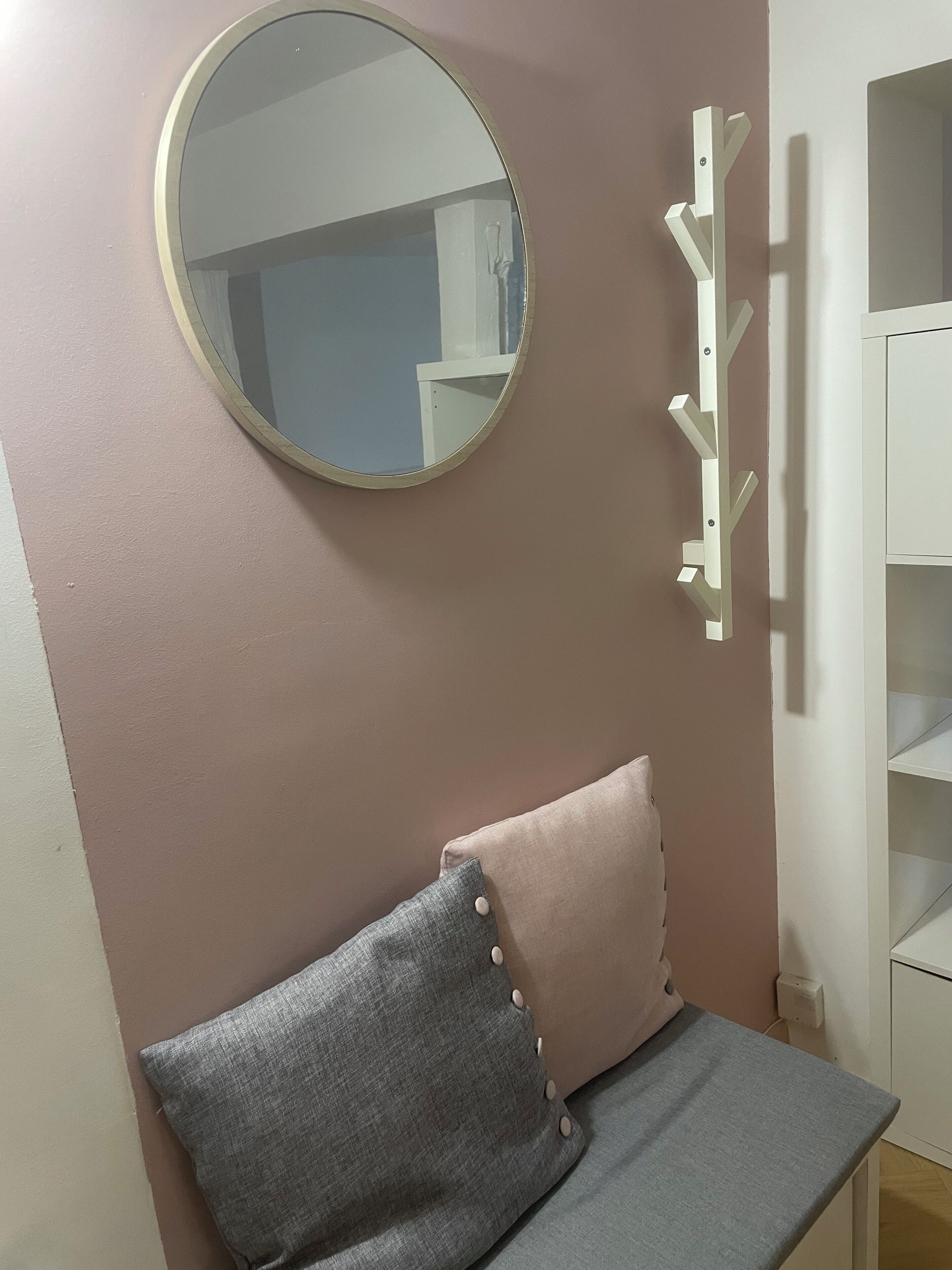 Une pièce avec un miroir rond, un canapé gris agrémenté d’un coussin rose et un porte-manteau blanc.