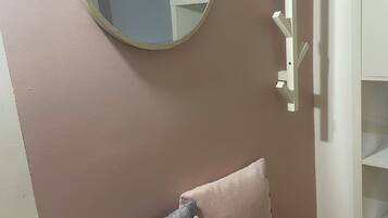 Une pièce avec un miroir rond, un canapé gris agrémenté d’un coussin rose et un porte-manteau blanc.