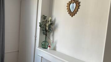 Ett rum med en vit garderob, ett litet runt bord med ett svart föremål på, en grön vas med växter och en hjärtformad spegel på väggen.