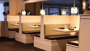 Une rangée de banquettes disposées comme dans un stand, avec des tables, chacune agrémentée d’une petite lampe de table et d’une plante posée sur le rebord de la fenêtre.