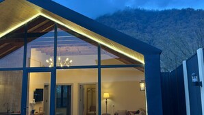 A modern house with large windows, a visible interior including a living room and a bathroom, and a mountainous backdrop. - Hg Barn House Gabala (Gabala)