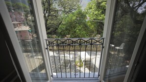 Un balcón con baranda de metal, suelo de madera y vistas a un jardín y edificios al fondo.