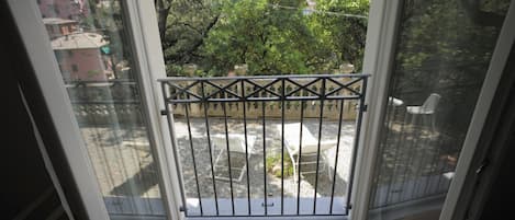 A balcony with a metal railing, a wooden floor, and a view of a garden and buildings in the background.