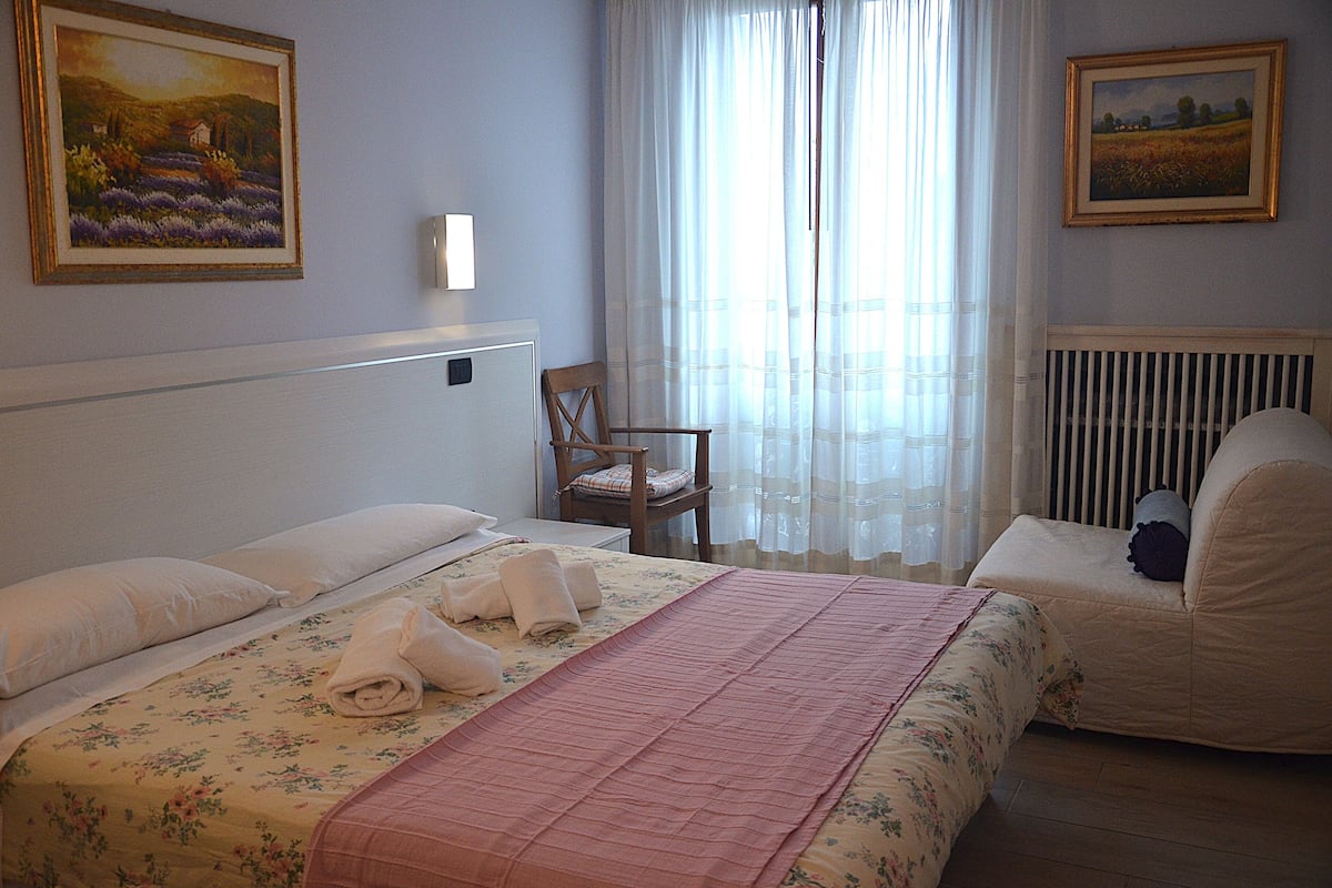 A hotel room with a bed, a chair, a radiator, and two paintings on the walls.