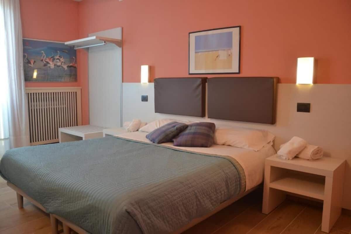 A bedroom with a bed, bedside tables, a radiator, a painting, and wall-mounted lights.