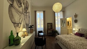 A hotel room with a large mural, a round chandelier, a bed, a desk with a lamp, and a view of the city through the window. - Haut de la Canebière Magnifique Appartement 4* (Marseille)