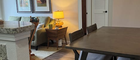 A dining area with a large wooden table and chairs, a small side table with a lamp, and framed pictures on the wall.