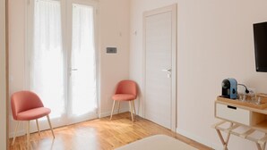 Una habitación con suelo de madera, dos sillas rosas, una puerta blanca, una pared blanca y una pequeña mesa de madera con una tetera y vasos.