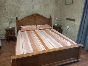 Ein Einzelbett mit Holzbettgestell und einem gestreiften Bettbezug. Zwei Nachttische, jeder mit einer Lampe. Die Wand im Raum ist aus Stein, auf der rechten Seite hängt ein Vorhang.